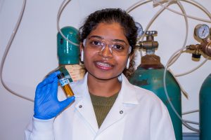Portrait of Thivani posing in a lab. She is wearing safety glasses, a lab coat, and gloves. 