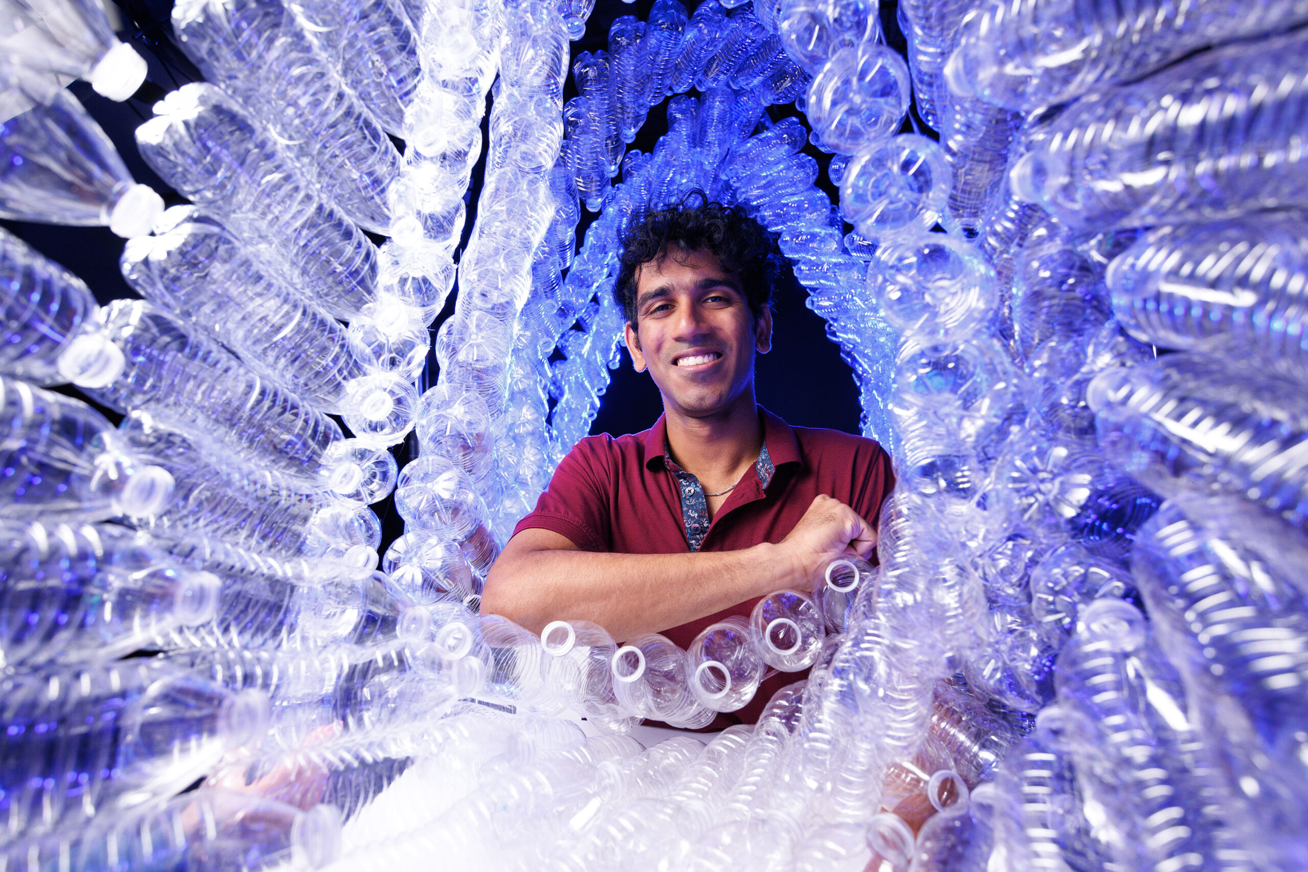 Dhananjay (DJ) Dileep poses for a portrait surrounded by plastic water bottles.